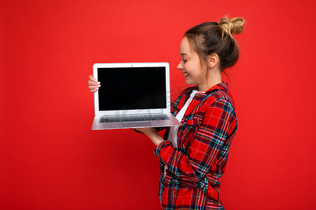 侧面照片：迷人漂亮的年轻女士手持上网本看着屏幕，身穿红色衬衫，与红墙背景隔离