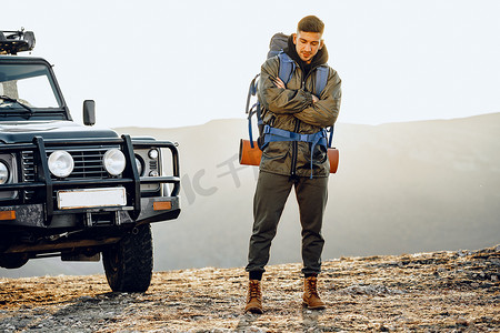 驾驶位摄影照片_一位身穿远足装备的年轻旅行者站在他的越野车旁的画像