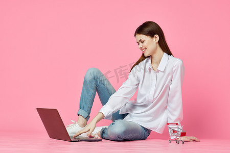 购物娱乐摄影照片_坐在地板上、带笔记本电脑购物娱乐粉红色背景的女人