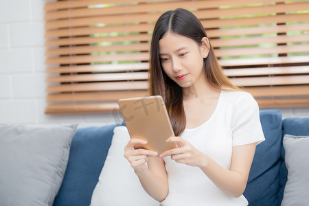 年轻迷人的亚洲女性在家里的沙发上使用浏览平板电脑休息，快乐的女孩坐在沙发上放松阅读家里的数字小工具、交流和生活方式概念。