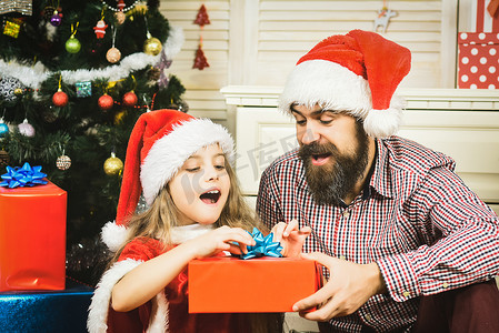 圣诞老人帽子摄影照片_快乐的父亲和女儿在圣诞树上带着戴着圣诞老人帽子的礼物。