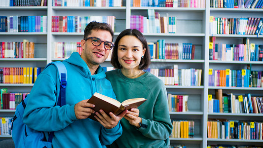 一位戴眼镜的年轻千禧一代学生和一位年轻漂亮的女学生正在读书，站在书架的背景下站在公共图书馆里准备考试。
