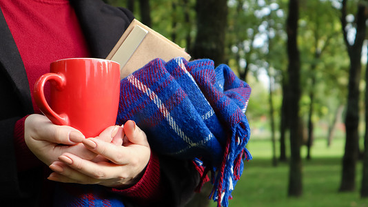 公园里一位穿着毛衣和黑色外套的女人手里拿着一杯红咖啡和一本书，上面有蓝色方格羊毛毯或格子花呢。