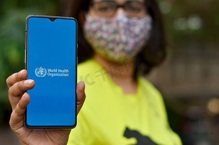 卫生负责摄影照片_美国，2020年。戴口罩的女孩在手机屏幕上显示，世界卫生组织（WHO）登陆页面和标志。联合国负责公共卫生的专门机构