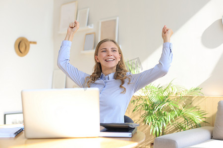 快乐的企业家女性坐在办公桌前阅读好消息，举起双手表达喜悦。