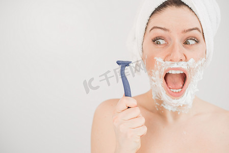 一个女人脸上涂着剃须泡沫的滑稽肖像，她拿着一把剃须刀，背景是白色的。