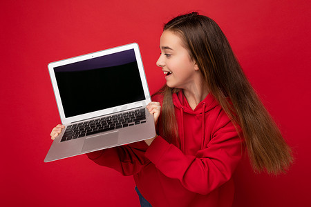 飞机侧面摄影照片_美丽快乐女孩的侧面照片，长发，身穿红色连帽衫，手持电脑笔记本电脑，看着红墙上隔开的上网本键盘和屏幕显示器