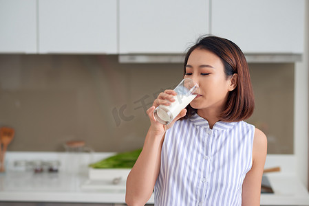 喝饮料的女孩摄影照片_年轻的亚洲女人站在厨房里喝牛奶的画像。