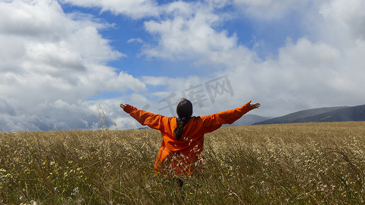 蓝天多云的日子里，身穿橙色夹克、双臂举起的年轻女子在麦田中间望着天空