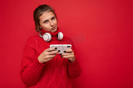app蓝摄影照片_穿着红色毛衣的迷人、不满的年轻深色女性在红色背景墙上被隔离，手持和使用手机在互联网上冲浪，戴着蓝牙耳机看着相机