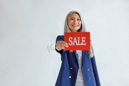 灰白摄影照片_头发灰白的美丽亚洲女性在浅色背景中展示带有“Sale”字样的红牌