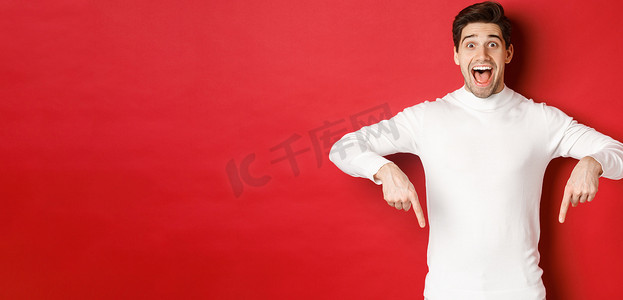 身穿白色毛衣的帅哥对圣诞广告反应兴奋，手指向下，展示标志，站在红色背景上