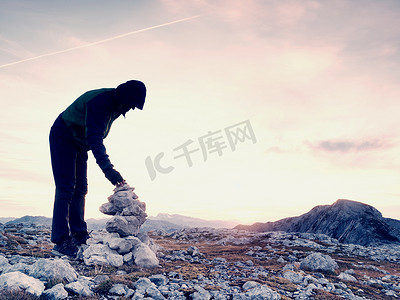 孤独的成年男子正在向金字塔堆放石头。阿尔卑斯山山顶，傍晚的阳光在地平线上。