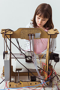 3d打印设计摄影照片_戴着 3d 打印百叶窗的可爱女孩正在看着她的 3d 打印机打印她的 3d 模型。