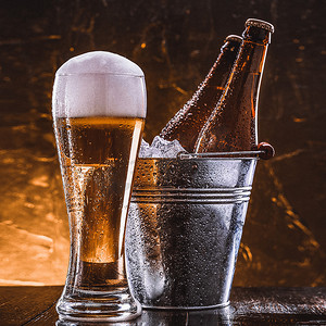 两瓶啤酒装在一个桶里，里面装着冰，还有一杯啤酒，泡沫丰富，紧挨着深色背景
