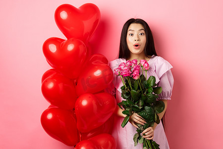 穿着裙子的亚洲女孩惊讶地站在情人节心形气球附近，对着镜头说哇，手捧情人送来的花束，浪漫的玫瑰约会，粉红色背景