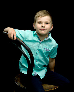 坐在凳子上的孩子摄影照片_微笑的小男孩坐在黑色背景的凳子上。