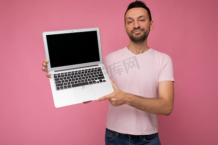 英俊的黑发男人微笑着拿着笔记本电脑，看着穿着 T 恤的相机，背景是孤立的粉红色