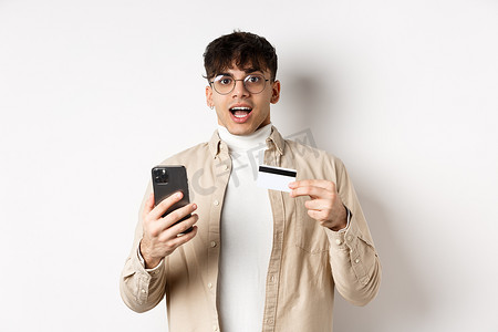 兴奋的年轻人在网上购物，拿着手机和塑料信用卡，在网上购物，站在白色背景上