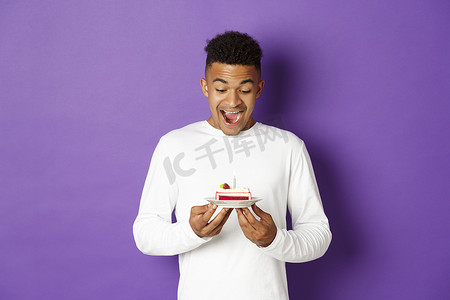 快乐的年轻非洲裔美国人庆祝生日的画像，看着生日蛋糕，微笑着许愿，站在紫色背景上