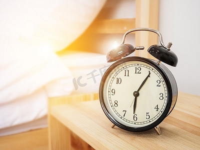 时钟表摄影照片_闹钟，叫醒时间概念：复古闹钟，早上六点过五分，在木床边桌上，配有白色床单和早晨阳光背景