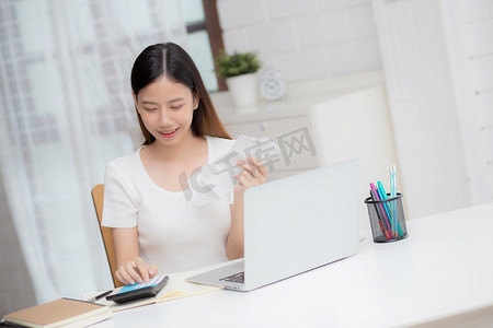 年轻的亚洲女性在家里用桌上的计算器计算财务家庭，女孩检查账单并成功节省家庭开支，债务清偿，税务和会计，商业概念。