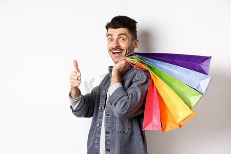 优惠商品摄影照片_兴奋的男性购物者肩上扛着购物袋，竖起大拇指，站在白色背景上推荐商店里的优惠商品