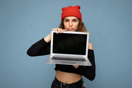 深色墙壁背景摄影照片_特写肖像的滑稽美丽的深色金发女人拿着笔记本电脑看着相机穿着黑色裁剪上衣和红色和橙色的 do-rag 隔离在浅蓝色的墙壁背景
