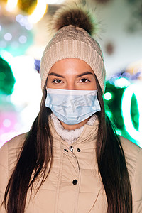 童话城摄影照片_戴着医用面罩的白人年轻女性站在装饰精美的圣诞城的垂直特写照片。