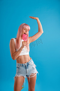 活泼的粉红色头发的女人玩得很开心，拿着梳子，在里面唱歌，同时站在蓝色的工作室背景中