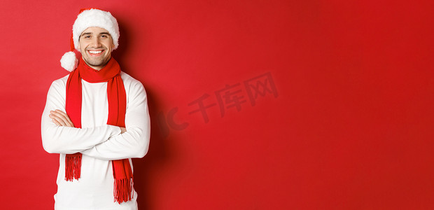 圣诞帽围巾摄影照片_帅气的白种人，长着鬃毛，戴着圣诞帽、围巾和白色毛衣，双臂交叉在胸前，笑得开心，站在红色背景下