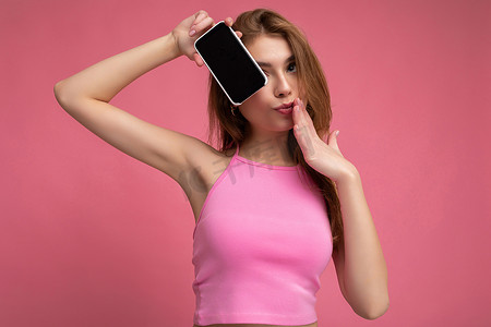 照片中，美丽积极的年轻金发女性身穿粉红色上衣，在粉红色背景中与世隔绝，手握空白空间，展示手机，显示屏为空屏，用于注视相机