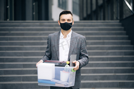 被解雇的悲伤的年轻商人戴着医用面具，带着装有个人物品的盒子离开办公室。
