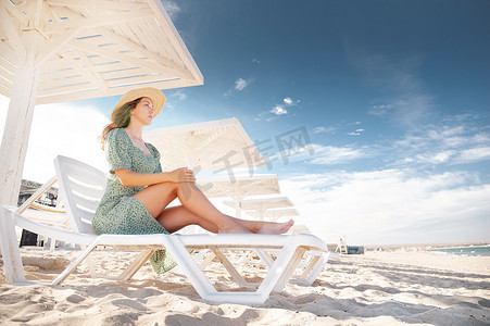 一位身穿绿色连衣裙的金发女郎坐在日光浴床上，在遮阳伞下看着海景。