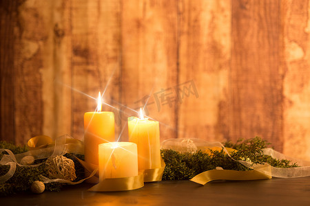 金色的缎带摄影照片_圣诞节概念的温暖：三根蜡烛点亮，十字屏幕星星在一张深色木桌上产生效果，桌子上有松枝、天然松果、金色缎带和白色欧根纱丝带