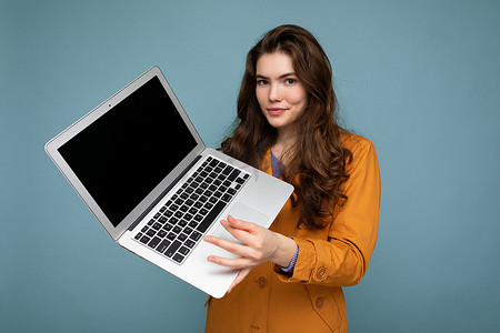 照片中，美丽严肃、快乐的年轻女子手持电脑笔记本电脑，看着身穿黄色夹克的相机，与蓝色墙壁背景隔离