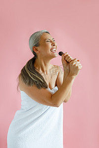 唱歌摄影照片_快乐的银发亚洲女士用刷子站在粉红色背景上唱歌