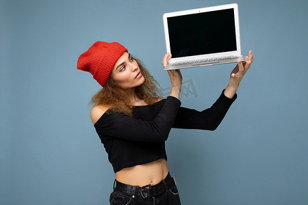美丽自信的深色金发卷发年轻女子的特写肖像，她手持笔记本电脑，看着上网本键盘和显示器，身穿黑色裁剪上衣和红色和橙色的 do-rag，与浅蓝色墙壁背景隔离