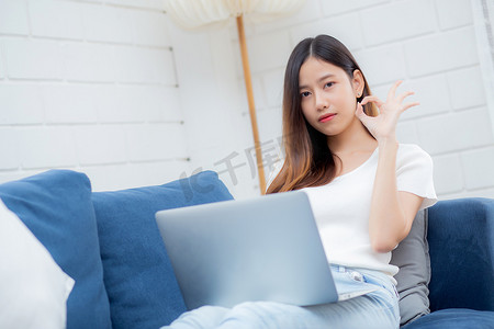 年轻的亚洲女商人微笑着在家里工作，带着笔记本电脑上网，在客厅的沙发上上网，自由职业的女孩在沙发上使用笔记本电脑，带着舒适、新常态、生活方式的概念。