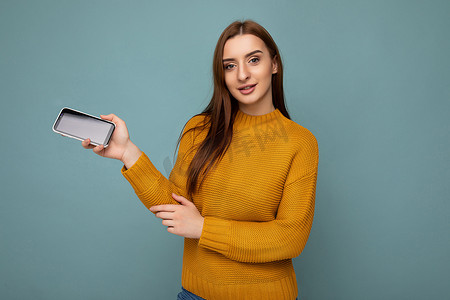 身穿橙色毛衣的漂亮积极年轻女性的照片，在蓝色背景中与世隔绝，手握空地，展示手机，空地用于模拟看相机