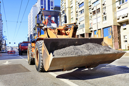 一名道路工人在道路维修期间用重型建筑推土机铲斗搬运碎石。