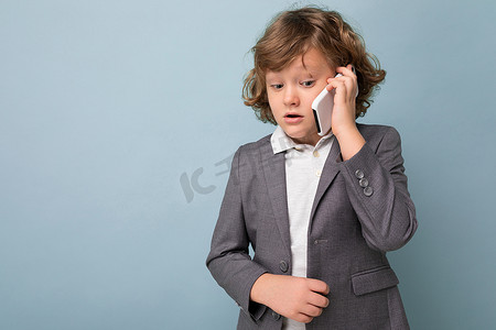 照片中，一头卷发、身穿灰色西装的英俊男孩拿着手机，在蓝色背景中被隔离，低头看着手机，与某人交谈