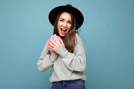 快乐积极微笑的年轻美丽迷人的黑发女人的肖像照片，带着真诚的情感穿着灰色毛衣和黑色帽子隔离在蓝色背景与复制空间和拿着粉红色的小猪盒。