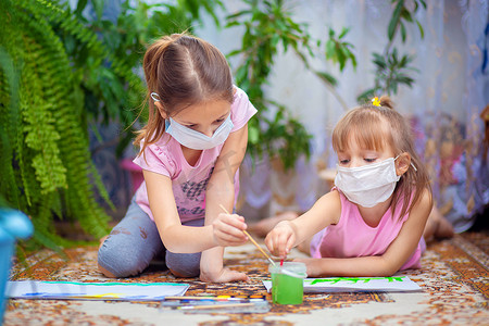 两个戴着防护口罩的女孩在家里或幼儿园的地板上涂油漆