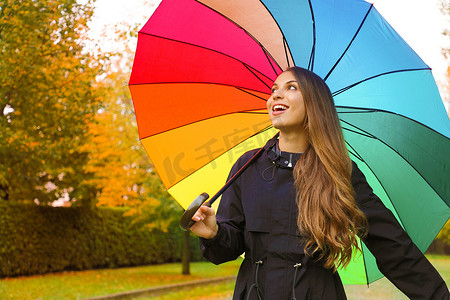 雨天在城市公园的彩虹伞下穿着深色雨衣的快乐女人的画像。