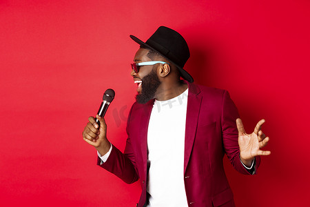 热情的黑人男歌手在红色背景下表演，对着麦克风唱歌，穿着派对服装，站在红色背景上