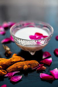 黑色闪亮表面上的牛奶面膜，由玻璃碗中的生牛奶、姜黄或 haldi 和 badam 以及全部原料组成。
