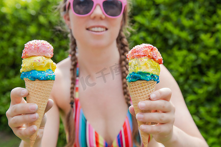 夏天，漂亮女孩拿着两块彩色冰淇淋，上面洒着彩虹色泳衣，粉色太阳镜和辫子，快乐假期，假期概念