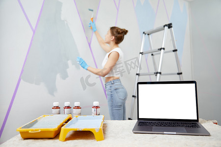 年轻女子用油漆滚筒和遮蔽胶带粉刷墙壁。