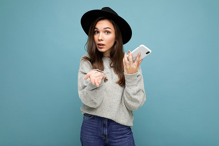 戴着黑帽和灰色毛衣的漂亮年轻女性拿着智能手机，看着背景中突显的相机，提出不满的褐发女性
